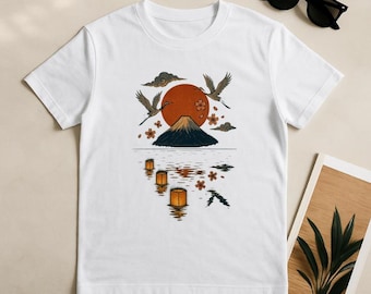 Pode incluir: Uma t-shirt branca de manga curta com uma impressão gráfica de uma montanha escura, um grande sol laranja, dois grous a voar e lanternas a flutuar na água. O design inclui detalhes de flores de cerejeira e nuvens escuras.