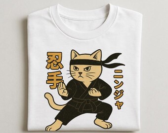 Könnte beinhalten: Weißes T-Shirt mit einer Cartoon-Ninja-Katze in schwarzer Uniform und Stirnband. Die Katze befindet sich in einer Kampfhaltung mit japanischem Text. Das Design verwendet die Farben Beige, Schwarz und Gold.