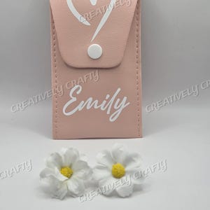 May include: A pale pink rectangular case featuring a white heart and exclamation mark design, and the name "Emily" in white. Two white daisy flowers with yellow centres are at the bottom. The case has a white button closure.