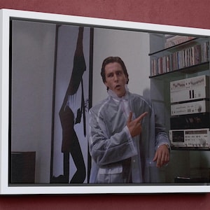 May include: Framed canvas art featuring a scene from a movie. The artwork depicts a man in a clear raincoat pointing to the side. A shelf with audio equipment and CDs is visible in the background. The frame is white.