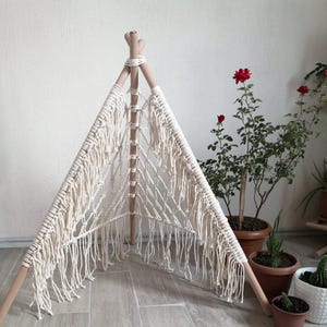 Op de afbeelding: Een crèmekleurige macramé tipi voor huisdieren met een houten frame. Het macramé heeft een geweven ontwerp met franjes. De tipi staat op een lichte houten vloer, met potplanten op de achtergrond. Geschatte hoogte: 100 cm.