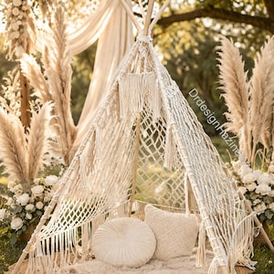 Op de afbeelding: Een bohemien-stijl tipi-tent gemaakt van natuurlijk hout en macrame-net, buiten opgesteld. De tent is versierd met crèmekleurige kussens, een rond kleed en pampasgras. De scène baadt in zacht zonlicht, wat een warme en uitnodigende sfeer creëert.