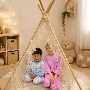 May include: Two children sit inside a beige teepee tent with a macrame fringe. The tent is made of wooden poles and sits on a round, beige rug. One child wears blue tie-dye pajamas, and the other wears pink tie-dye pajamas. The text "drdesignHazineleri" is at the bottom.
