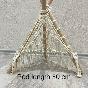 Op de afbeelding: Een kleine, handgemaakte tipi-achtige structuur met een macramé net en houten palen. De palen in natuurlijke tinten zijn in een driehoekige vorm gerangschikt, waarbij het macramé een geweven voorkant creëert. De tekst op de afbeelding luidt: Staaf lengte 50 cm.