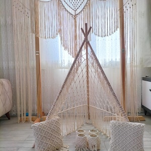 Op de afbeelding: Een macramé speeltent in tipi-stijl met een houten frame, versierd met crèmekleurige geweven details. Twee bijpassende kussens en vier kleine macramé plantenpot covers staan voor de tent. De achtergrond bevat een macramé wandkleed en een transparant gordijn.