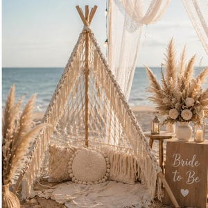 May include: A beach setup with a macrame teepee, pillows, and a rug. Dried pampas grass and flowers decorate the scene. A wooden sign reads "Bride to Be". String lights and sheer fabric add to the ambiance.