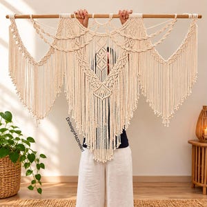 Op de afbeelding: Een grote gebroken witte macramé wandhanger opgehangen aan een horizontale houten roede. Het stuk bevat ingewikkelde knooppatronen, gevlochten secties en lange verticale franjes die eindigen in kleine knopen. De tekst DrdesignHazineleri is zichtbaar.
