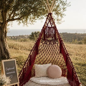 May include: A burgundy macrame teepee with a cream-colored rug, pillows, and a small chalkboard sign that reads "Captured memories". The teepee is set up outdoors with a tree and a view of the ocean in the background.