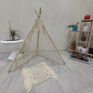 Op de afbeelding: Een crèmekleurige macramé tipi met houten palen en een bijpassend kleed. De tipi staat binnen, met een houten plantenstandaard en diverse potplanten op de achtergrond. Het kleed is rechthoekig met een gefranjerde rand.
