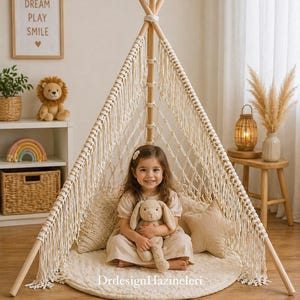 May include: A child sits inside a beige macrame teepee tent, holding a stuffed bunny. The tent has wooden poles and a cream-colored mat. A framed print with the words "DREAM PLAY SMILE" hangs on the wall.