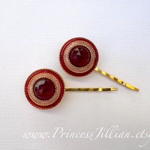 Puede incluir: Dos horquillas decorativas con un centro de joya roja, rodeadas de detalles rosas y dorados. Las horquillas tienen una base dorada y están sobre un fondo blanco. Se ve la dirección del sitio web www.PrincessJillian.etsy.com.