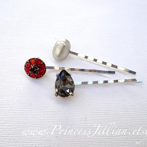 Può includere: Un assortimento di forcine decorative. Una forcina presenta un design circolare rosso e nero, un'altra ha un gioiello grigio scuro a forma di pera e una terza ha una forma ovale argentata. Le forcine hanno una finitura argentata.
