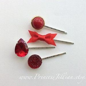 May include: Four red hair clips with different embellishments. One clip has a round red gem, another has a teardrop-shaped red gem, another has a red bow, and the last has a round red rhinestone.