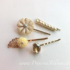 May include: Set of five gold hair clips with decorative accents. One clip features a white flower with a rhinestone center, another has a white flower with a pearl center, and another has a gold filigree design. The remaining two clips have a single rhinestone and a row of pearls.