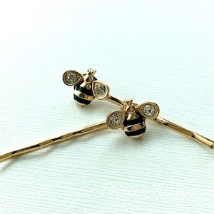 以下が含まれることがあります： 黒と白のストライプの体にクリアなラインストーンのアクセントが付いた、ゴールドの蜂のヘアクリップ2個