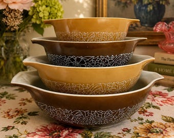 May include: A stack of four nesting mixing bowls with handles, alternating between golden yellow and dark brown colors. Each bowl features a white line-art floral pattern around the exterior. The bowls are arranged from smallest to largest.
