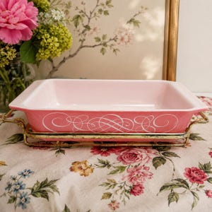 May include: A rectangular pink ceramic baking dish with a white scrolled filigree pattern on the exterior sides. The dish is set within a gold-toned metal wire frame that features two side handles for lifting and carrying the vessel.
