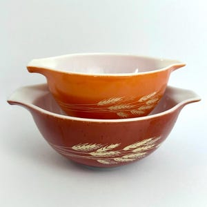 May include: Two vintage Pyrex mixing bowls. The larger bowl is a deep reddish-brown color, and the smaller bowl is orange. Both bowls have a white interior and handles. A wheat pattern in a cream color decorates the exterior of the bowls.