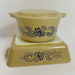 May include: Two-piece vintage tan casserole dish set with a speckled finish and dark blue floral design. The round casserole dish has a lid and is stacked on a square baking dish. Both pieces have handles.