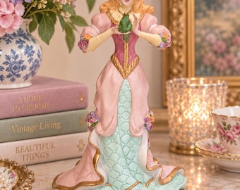 May include: A porcelain figurine of a woman with long blonde hair wearing a pink and light green gown with gold trim, holding a small green frog in her hands. The dress features floral accents at the shoulders and waist with a quilted pattern.