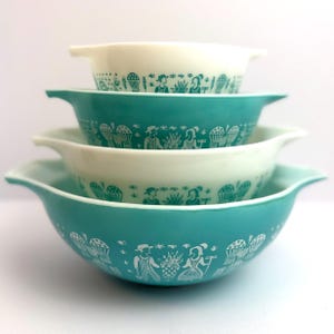 May include: A stack of four vintage Pyrex mixing bowls in shades of teal and cream. Each bowl features a white, stylized design of people, baskets, and plants. The bowls have a classic, rounded shape with handles.