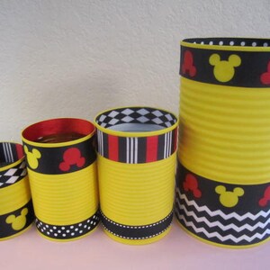 K&ouml;nnte beinhalten: Vier gelbe Dosen, verziert mit schwarz-wei&szlig;en Chevron-, Punkt- und Karomustern sowie rotem und schwarzem Band mit Mickey-Maus-Ohren.