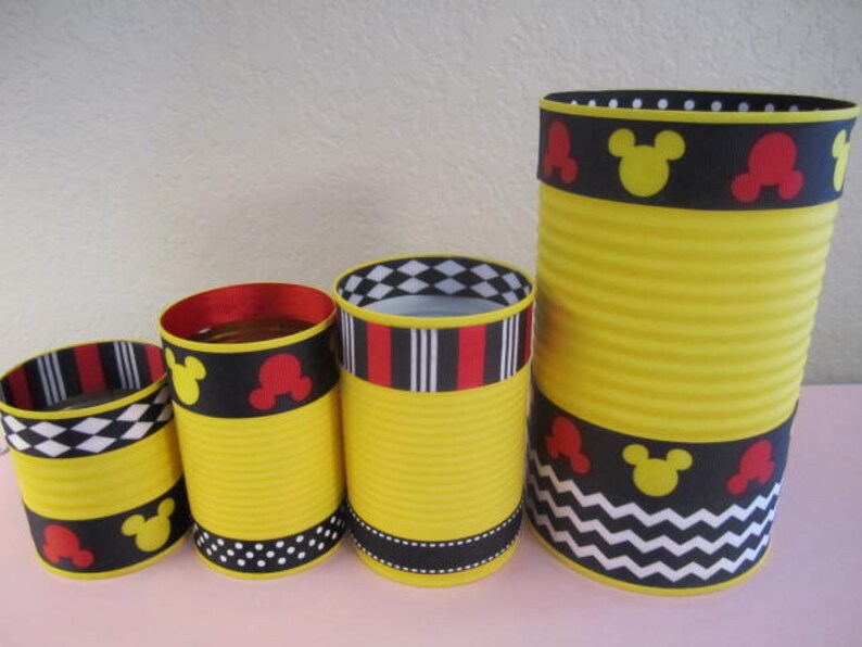 K&ouml;nnte beinhalten: Vier gelbe Dosen, verziert mit schwarz-wei&szlig;en Chevron-, Punkt- und Karomustern sowie rotem und schwarzem Band mit Mickey-Maus-Ohren.