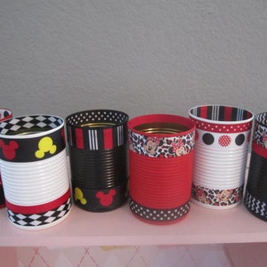 May include: Six decorated metal cans with red, black, and white polka dot, chevron, and Mickey Mouse themed ribbon. The cans are arranged in a row on a pink shelf.