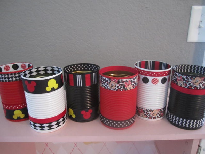 May include: Six decorated metal cans with red, black, and white polka dot, chevron, and Mickey Mouse themed ribbon. The cans are arranged in a row on a pink shelf.
