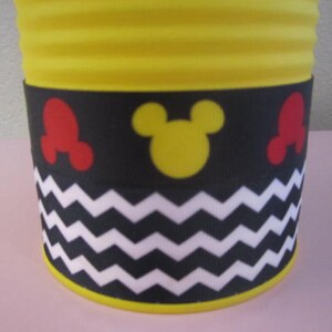 K&ouml;nnte beinhalten: Gelber zylindrischer Beh&auml;lter mit einem schwarz-wei&szlig;en Zickzackmuster, roten und gelben Mickey-Mouse-Ohren und einem gelben Mickey-Mouse-Kopf.