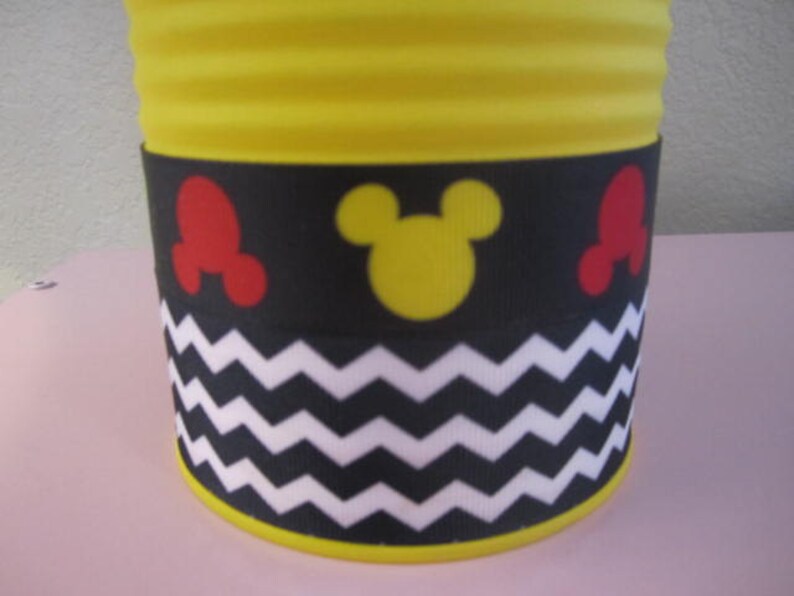 K&ouml;nnte beinhalten: Gelber zylindrischer Beh&auml;lter mit einem schwarz-wei&szlig;en Zickzackmuster, roten und gelben Mickey-Mouse-Ohren und einem gelben Mickey-Mouse-Kopf.