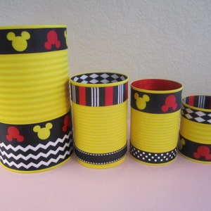 K&ouml;nnte beinhalten: Vier gelbe zylindrische Beh&auml;lter mit schwarzem, rotem und wei&szlig;em gemustertem Band um die Oberseite. Die Beh&auml;lter sind mit Mickey-Maus-Ohren und -Gesichtern verziert.
