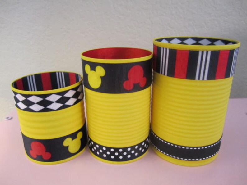 K&ouml;nnte beinhalten: Drei gelbe zylindrische Beh&auml;lter mit schwarzem, rotem und wei&szlig;em gemustertem Band. Die Beh&auml;lter haben ein Mickey-Mouse-Motiv mit schwarzen Mickey-Mouse-Ohren und roten Mickey-Mouse-K&ouml;pfen.