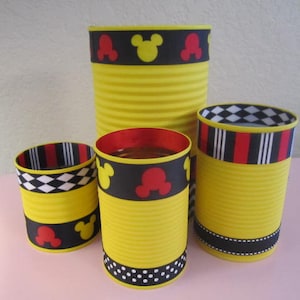 K&ouml;nnte beinhalten: Vier gelbe Dosen, die mit schwarzem und rotem Mickey-Mouse-Band verziert sind. Die Dosen haben unterschiedliche Gr&ouml;&szlig;en und Designs.