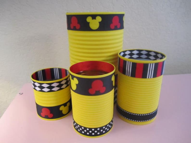 K&ouml;nnte beinhalten: Vier gelbe Dosen, die mit schwarzem und rotem Mickey-Mouse-Band verziert sind. Die Dosen haben unterschiedliche Gr&ouml;&szlig;en und Designs.