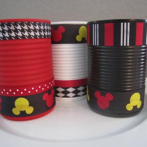 May include: Three empty cans decorated with Mickey Mouse themed ribbon. The cans are red, white, and black. The ribbon features black and white houndstooth, red and white stripes, and black with red and yellow Mickey Mouse heads.