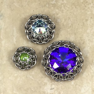 May include: Three silver chainmaille rings with different coloured gemstones in the centre. The rings are on a light brown surface.