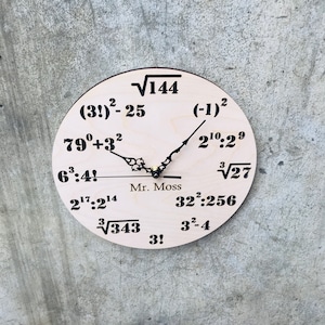 Puede incluir: Un reloj de madera con un dise&ntilde;o &uacute;nico que presenta ecuaciones matem&aacute;ticas en lugar de n&uacute;meros. La esfera del reloj es de color marr&oacute;n claro con texto negro y una aguja de reloj negra. El texto en la esfera del reloj incluye ecuaciones como "(3!)-25", "79&deg;+32", "6:4!", "217:214", "&radic;343", "3!", "(-1)2", "210:29", "&radic;27", "322:256", "32-4" y "&radic;144". El reloj tambi&eacute;n incluye el texto "Mr. Moss".
