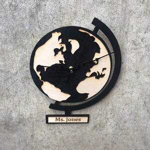 May include: Black wooden globe clock with a white outline of the continents. The clock has a black stand and a nameplate that reads "Ms. Jones".
