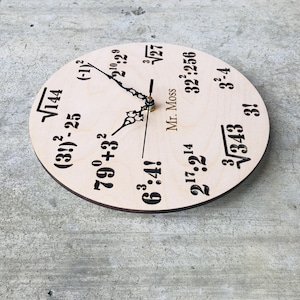 Puede incluir: Un reloj de madera con ecuaciones matem&aacute;ticas en lugar de n&uacute;meros. La esfera del reloj es de color marr&oacute;n claro con texto negro. Las ecuaciones incluyen ra&iacute;ces cuadradas, exponentes y factoriales. Las manecillas del reloj son negras y doradas. El texto "Mr. Moss" est&aacute; impreso en la esfera del reloj.