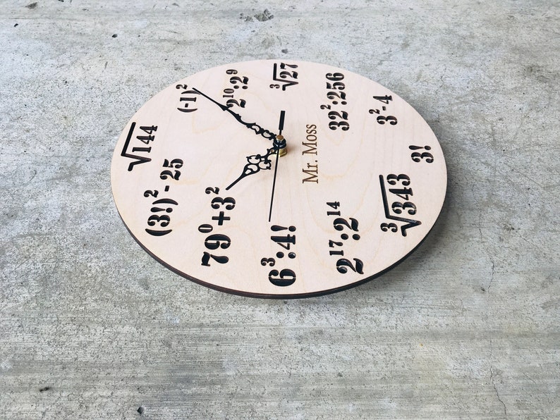 Puede incluir: Un reloj de madera con ecuaciones matem&aacute;ticas en lugar de n&uacute;meros. La esfera del reloj es de color marr&oacute;n claro con texto negro. Las ecuaciones incluyen ra&iacute;ces cuadradas, exponentes y factoriales. Las manecillas del reloj son negras y doradas. El texto "Mr. Moss" est&aacute; impreso en la esfera del reloj.