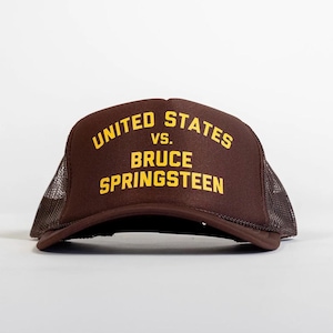 Puede incluir: Gorra de camionero marrón con el texto "UNITED STATES vs. BRUCE SPRINGSTEEN" en letras amarillas. La gorra tiene una parte trasera de malla y una visera curva. Estilo clásico de camionero.