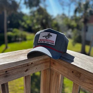 Könnte beinhalten: Eine Baseballkappe in Jeansoptik mit einem rechteckigen Aufnäher mit der Aufschrift "Winchester Rifles and Shotguns" und einer Grafik. Die Kappe hat eine rote Kordel und einen gebogenen Schirm. Der Hut liegt auf einem Holzgeländer.