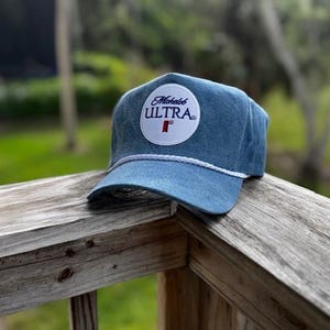 May include: A blue denim baseball cap with a white rope brim and a circular patch. The patch features the words "Michelob ULTRA" in a white circle with a red "T" below the text. The hat is resting on a wooden surface.