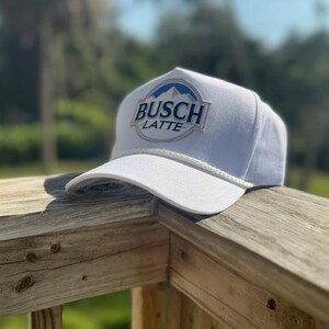 Peut inclure: Casquette de baseball blanche avec le texte "BUSCH LATTE" en bleu et blanc. Le chapeau a une visière incurvée et un détail de corde blanche. La casquette est posée sur une balustrade en bois.