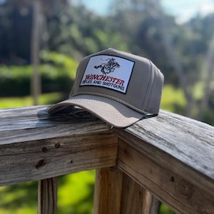 May include: A tan baseball cap with a rectangular patch featuring the Winchester Rifles and Shotguns logo. The logo includes a cowboy riding a horse. The cap is resting on a wooden railing.