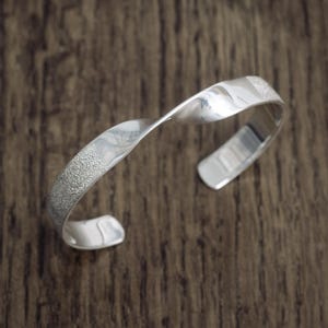 May include: A silver-toned cuff bracelet featuring a twisted design. The metal surface alternates between a smooth, reflective polished finish and a textured, granular matte finish, creating a distinct contrast along the length of the band.