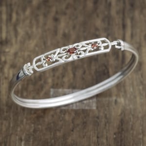 Könnte beinhalten: Ein silberner Armreif mit einem zentralen, rechteckigen Filigran-Element, in das drei kleine, oval geschliffene, tiefrote Granat-Edelsteine in einer Reihe eingefasst sind. Das Metallband ist glatt poliert und mit Scharnieren verbunden.