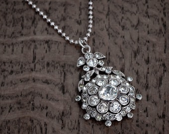 May include: A silver pendant necklace with a floral design. The pendant features a large, round, clear stone surrounded by smaller, clear stones. The necklace chain is a delicate ball chain.