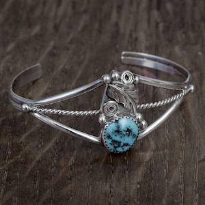 Peut inclure: Un bracelet manchette en argent orné d'une pierre turquoise ovale centrale avec une matrice noire, agrémenté d'un motif de feuille en argent, d'un design en spirale et de petites sphères, sur un bracelet en fil torsadé et fil lisse.